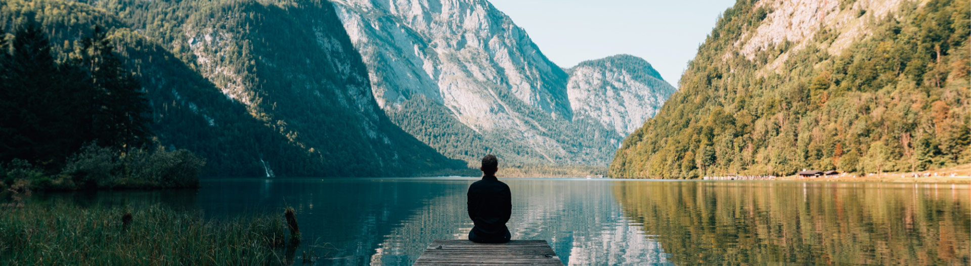 Photo of a person sitting at the end of a dock looking thoughtfully out at the distance. The dock extends into a lake in the middle of mountains.